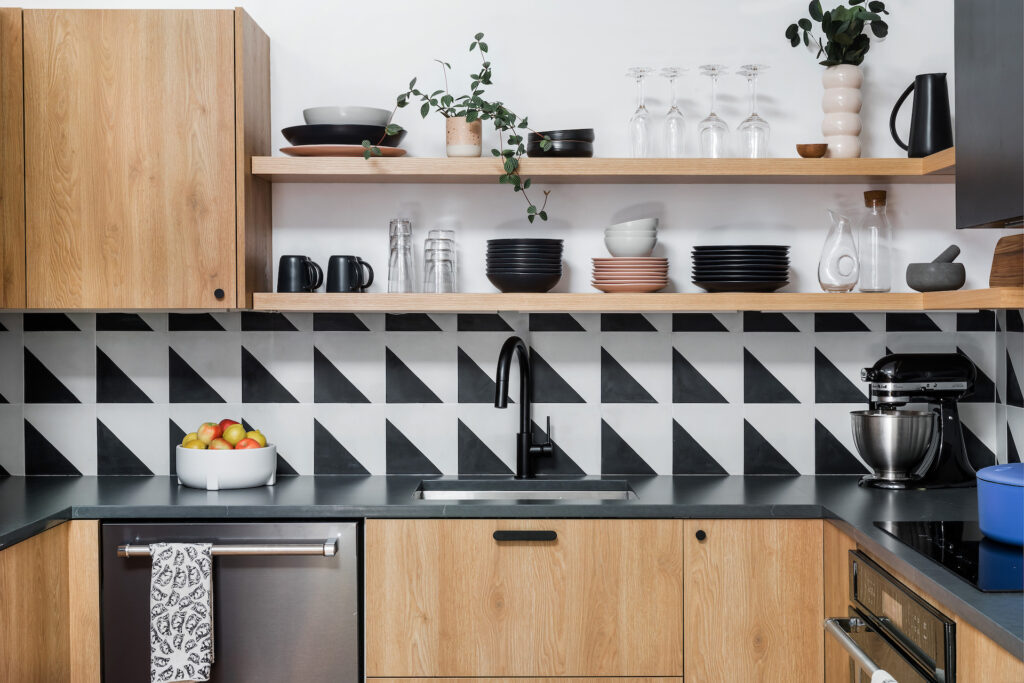 full view of the kitchen with wooden cabinets, open shelving, black countertops and hardware, and a black and white triangle backsplash.